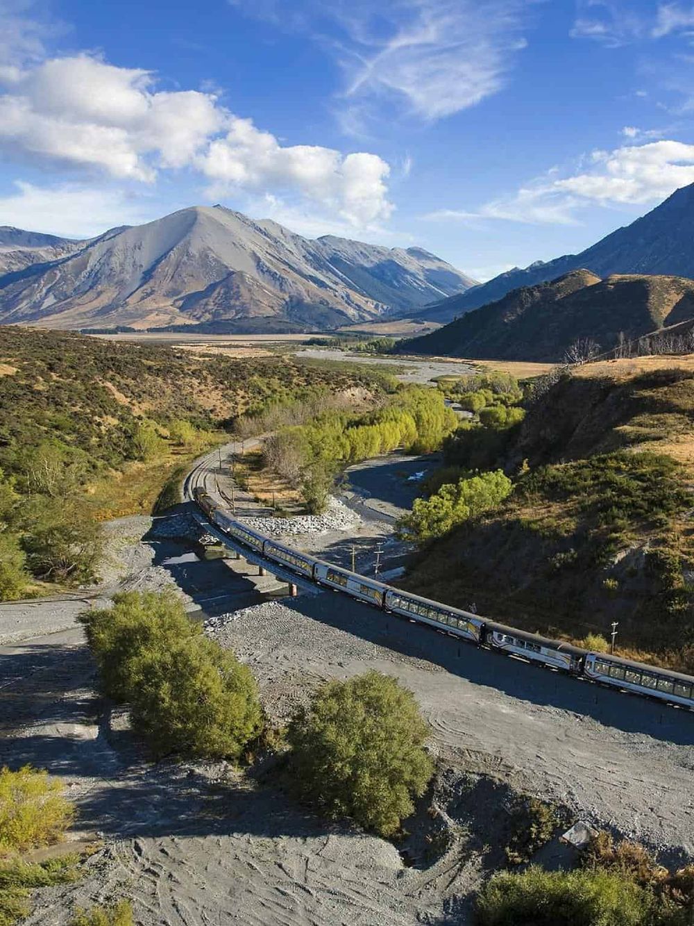 Scenic mountain landscape with a train crossing a river valley, lush greenery, and clear blue skies.