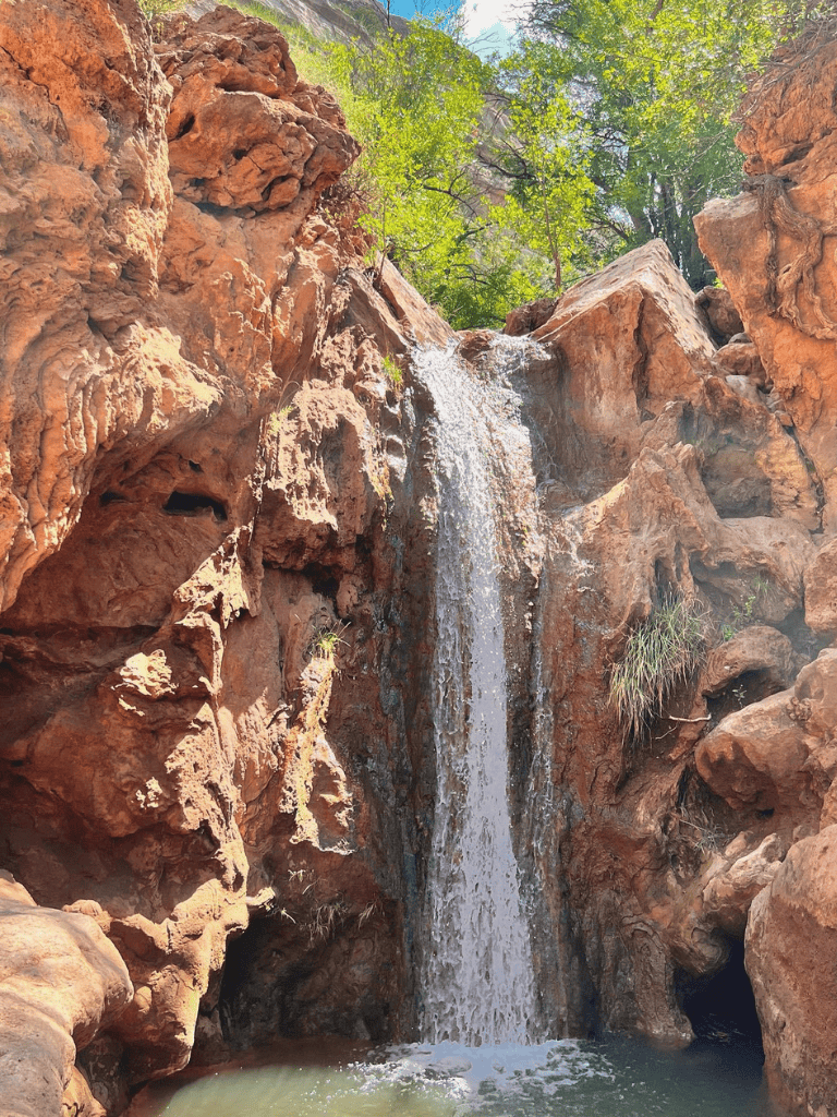 Serene desert waterfall cascading over red rocks with lush greenery, perfect for nature and adventure seekers.