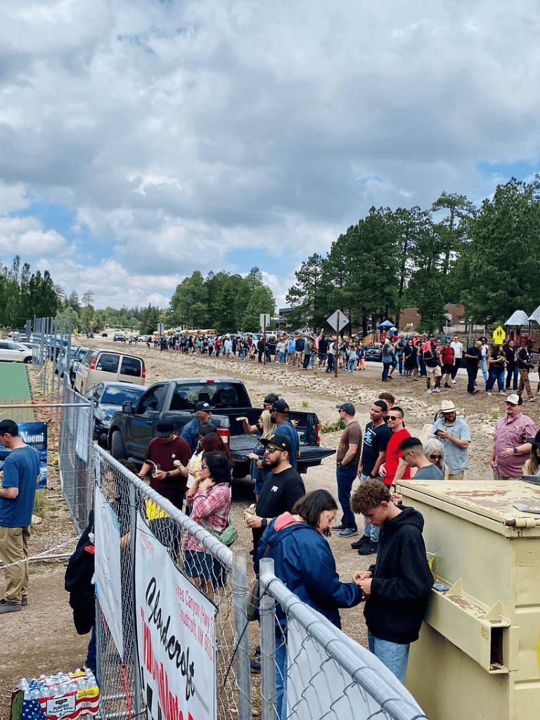 Crowd gathered at outdoor event with trees and cloudy sky, featuring long lines and vendor booths.