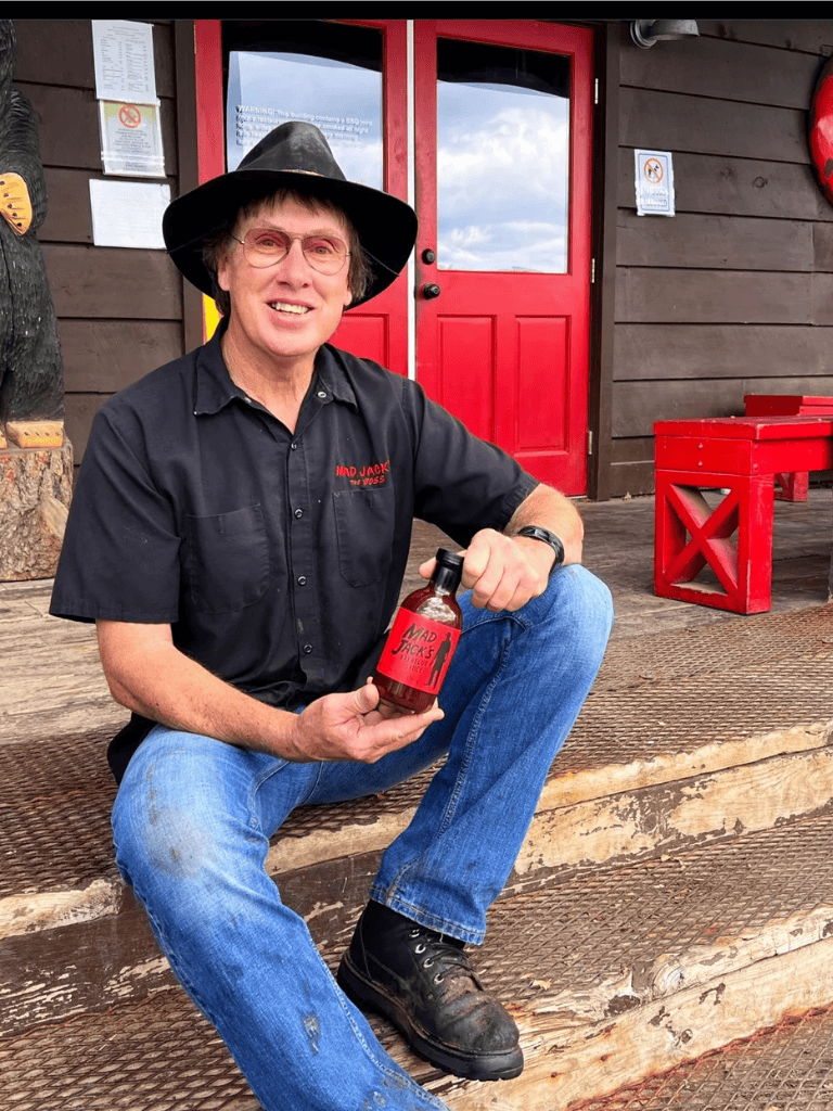 Mad Jack holding a hot sauce bottle outside a rustic building with red doors.