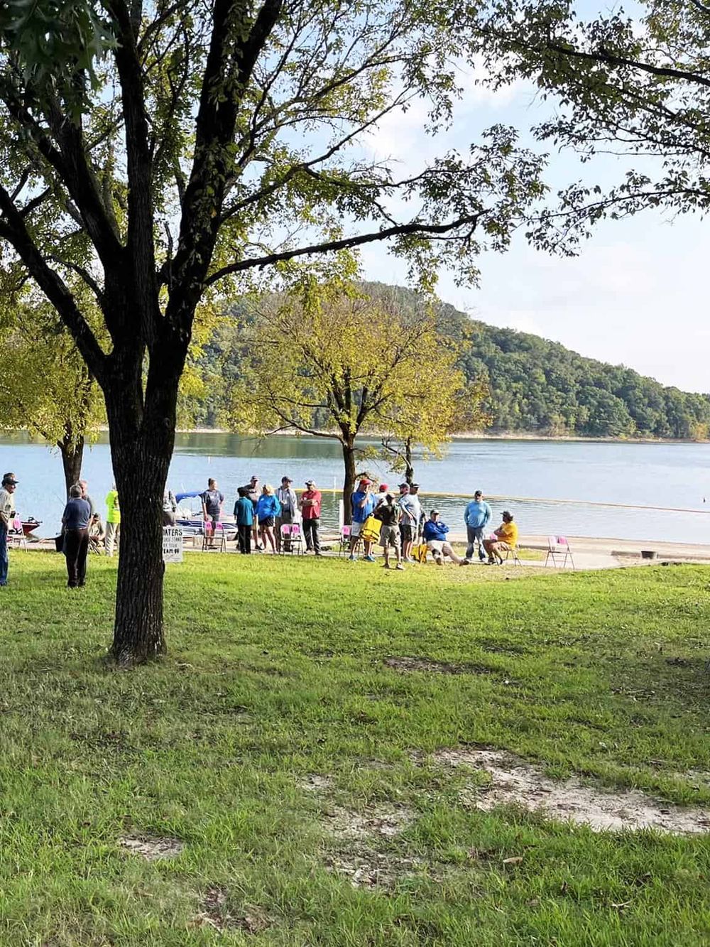 People gathered at a scenic lakeside park for an outdoor event or community gathering. Lush green trees and calm water create a peaceful setting.