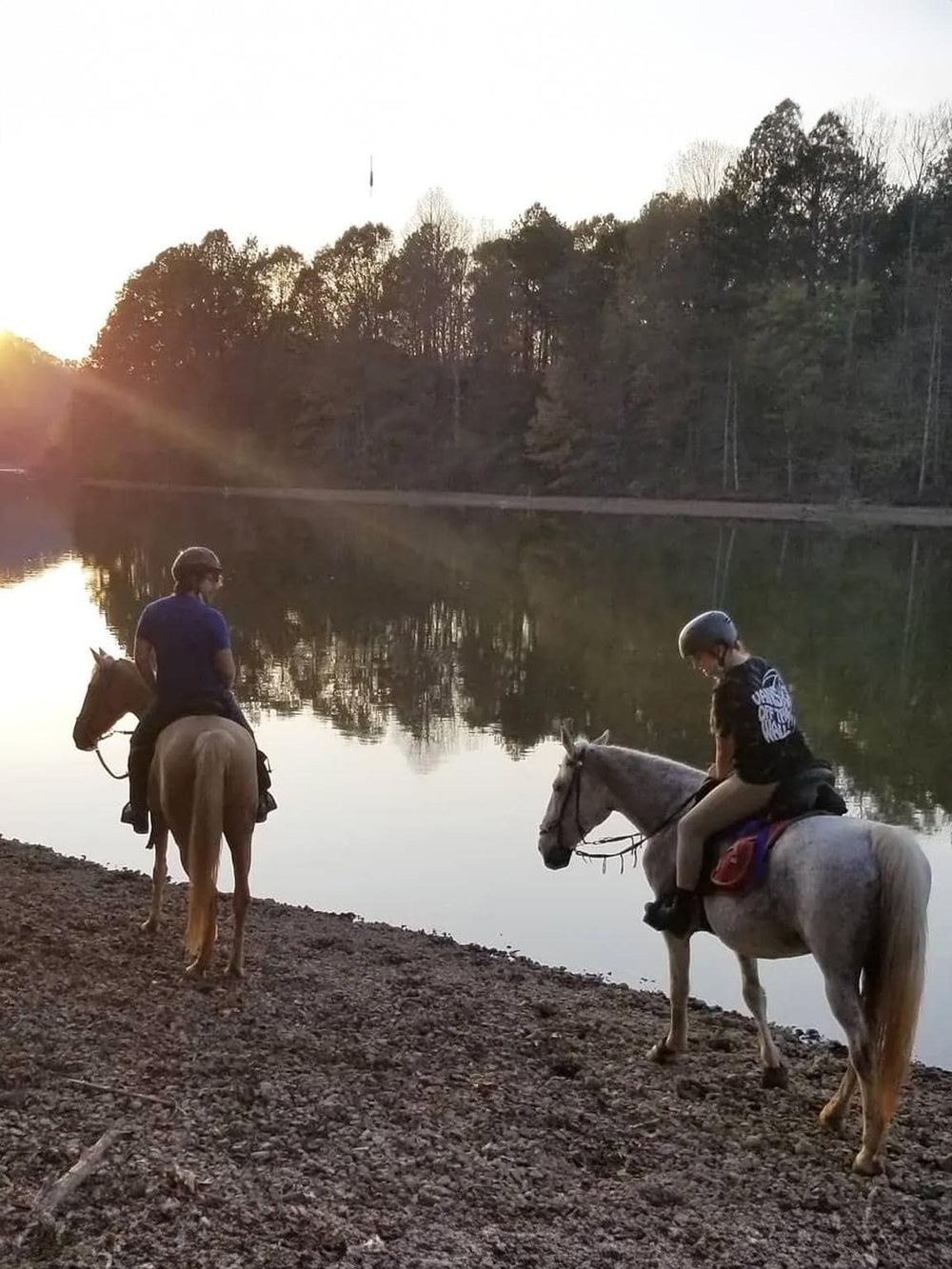 User image description: Two people horseback riding along a peaceful river at sunset, enjoying outdoor adventures with QuestForDirections.