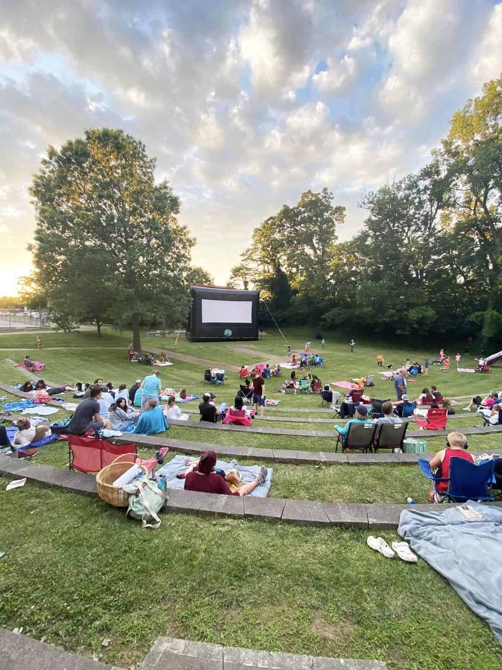 Outdoor movie night at a park featuring a large inflatable screen with people sitting on blankets and chairs on a grassy amphitheater.