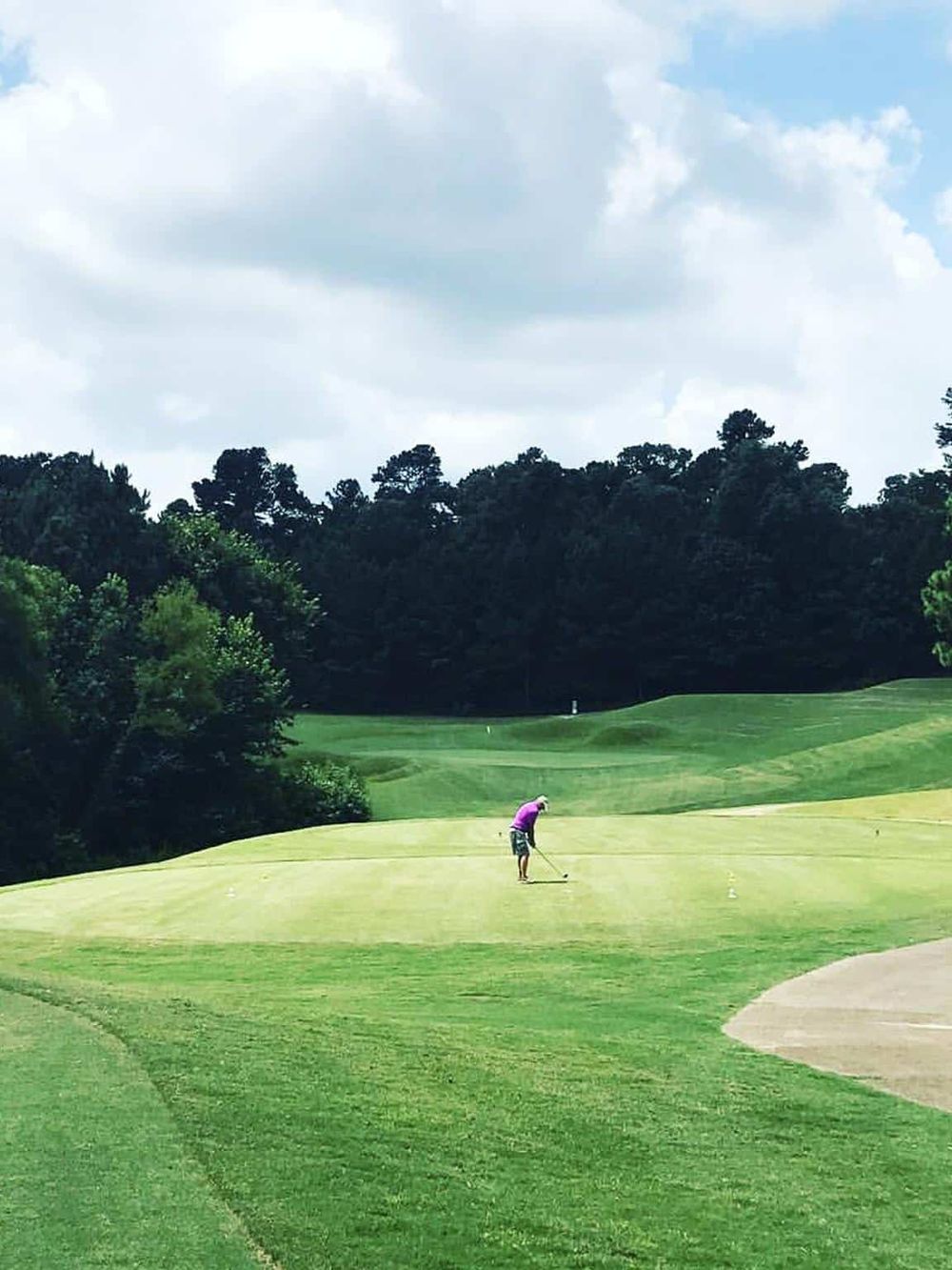 Golf course with a player preparing to putt, lush green fairways, trees and cloudy sky, outdoor sports and recreational activities.