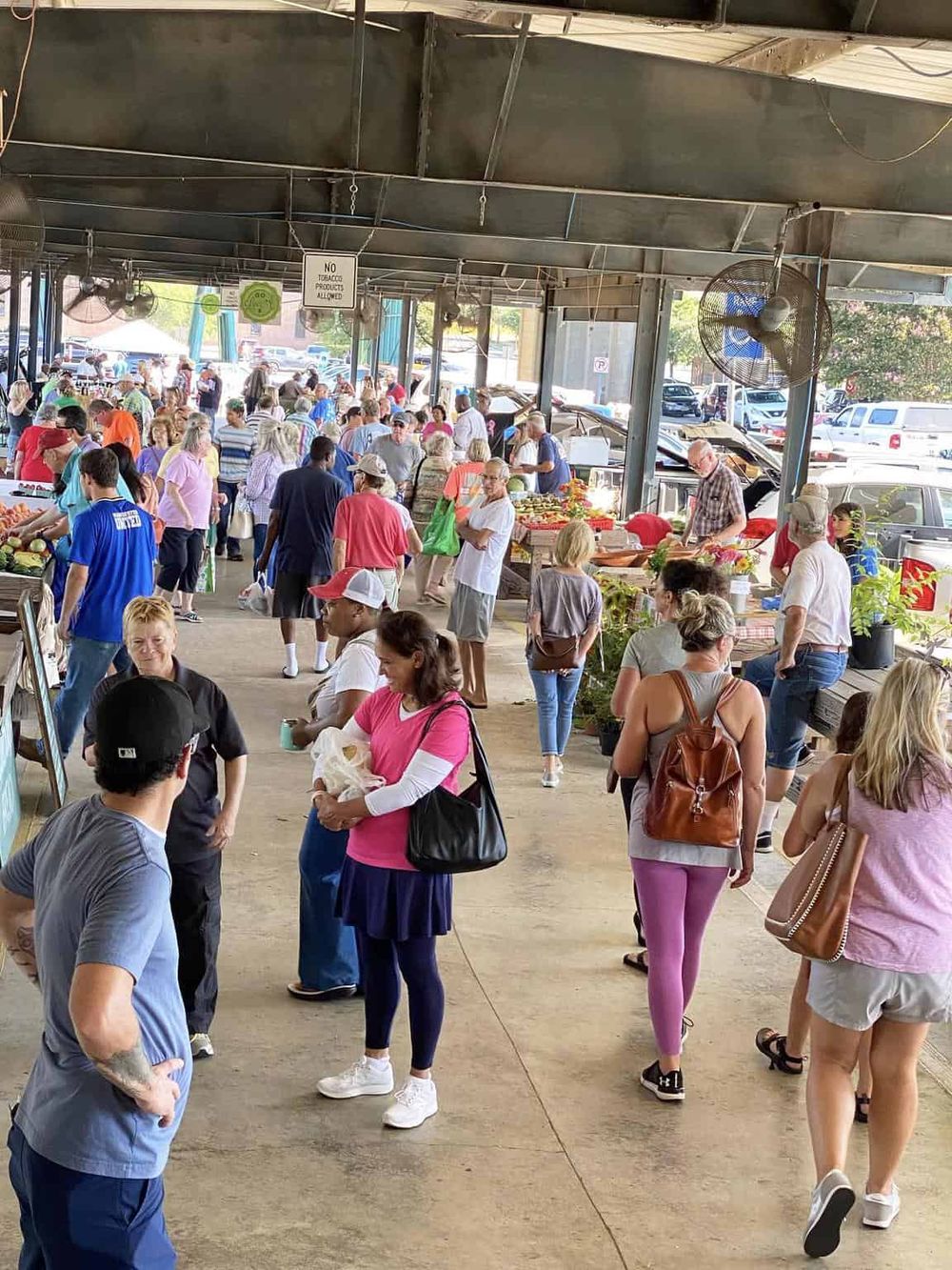 Crowded outdoor farmers market at QuestForDirections, with diverse shoppers buying fresh produce under a covered pavilion.