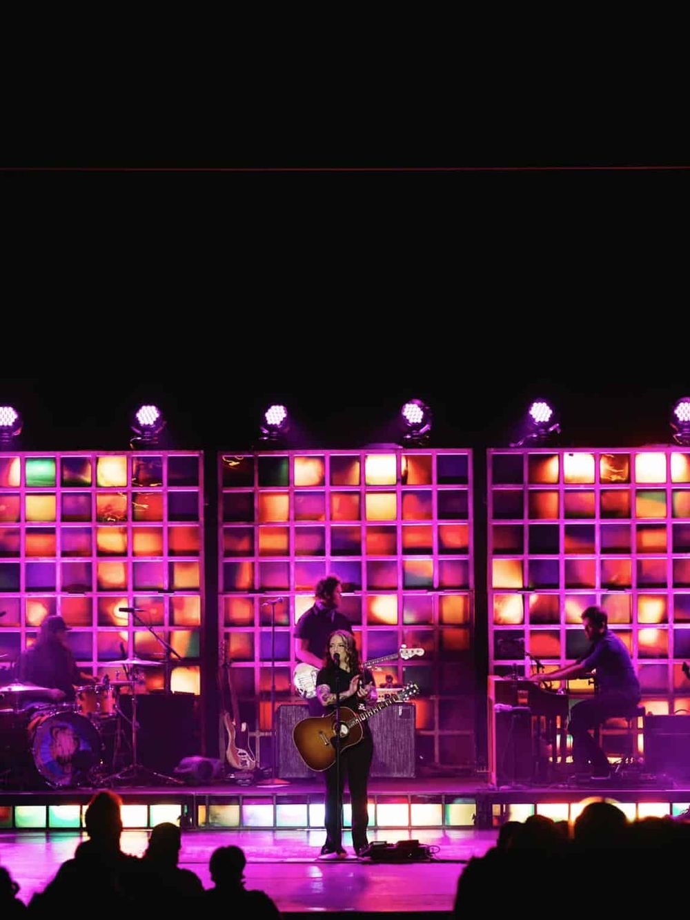 Vibrant live music performance featuring a female singer with guitar, band members, colorful stage lighting, and audience.