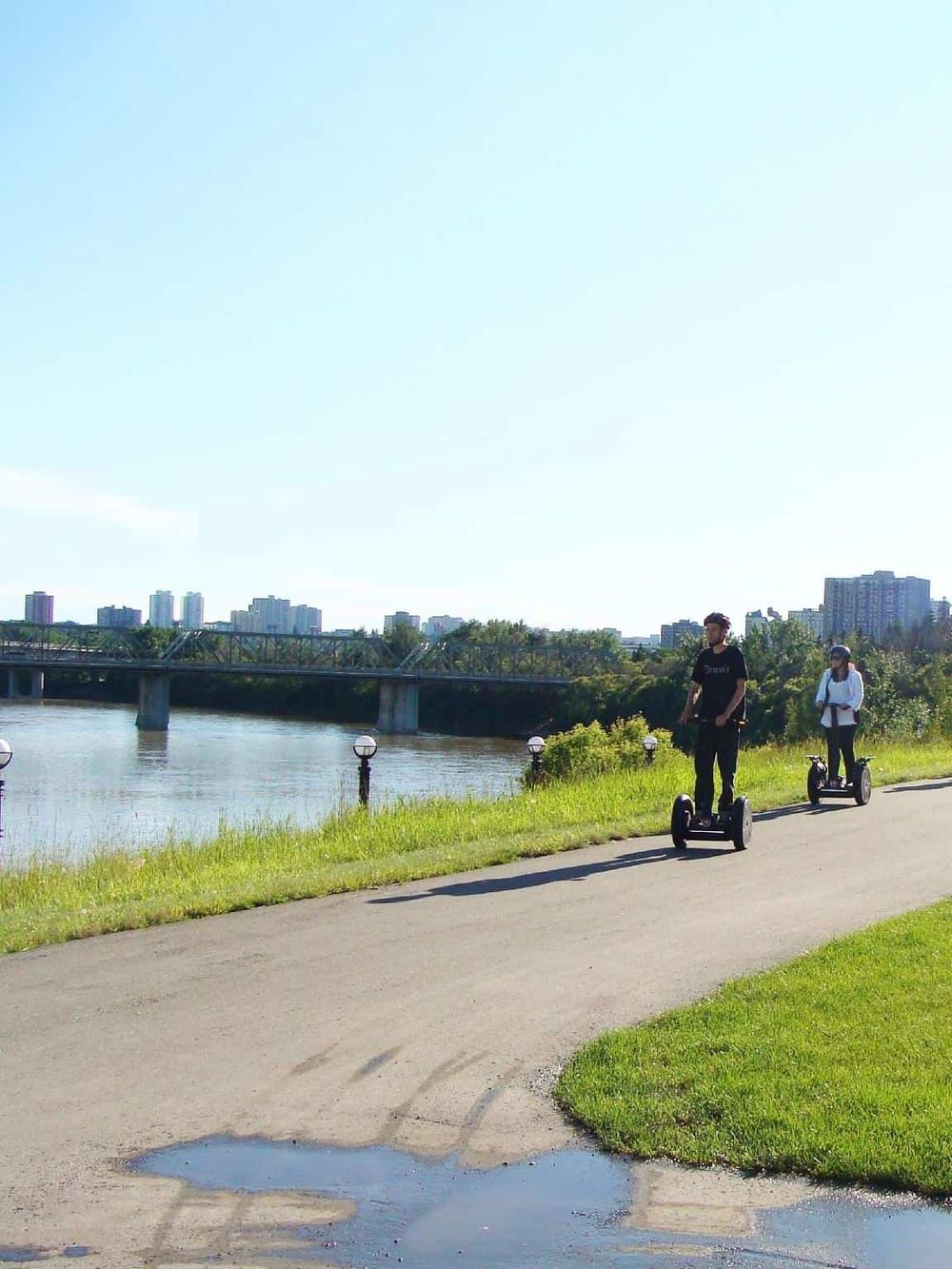 Riders on electric scooters enjoying a scenic park by the river with city skyline in background.