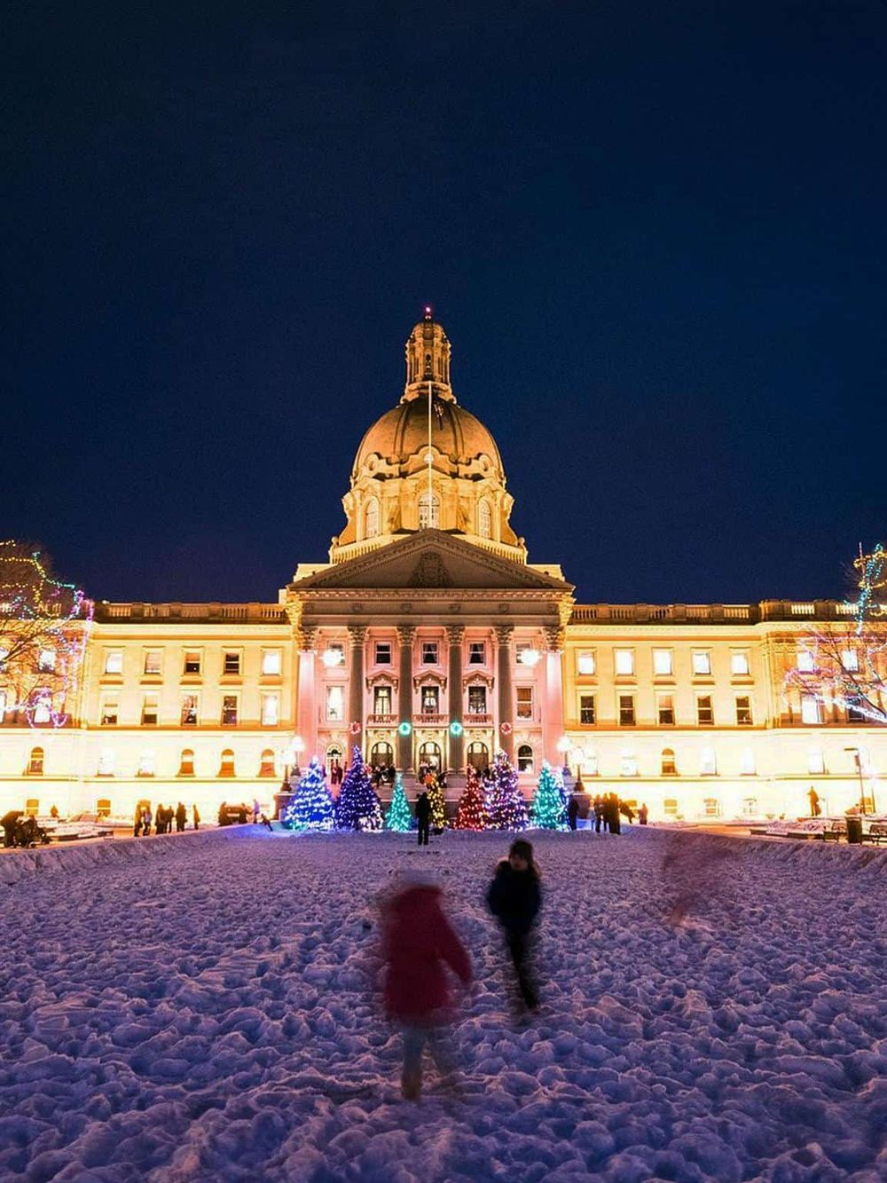 Stately Capitol building illuminated at night with festive holiday lights in snowy surroundings.
