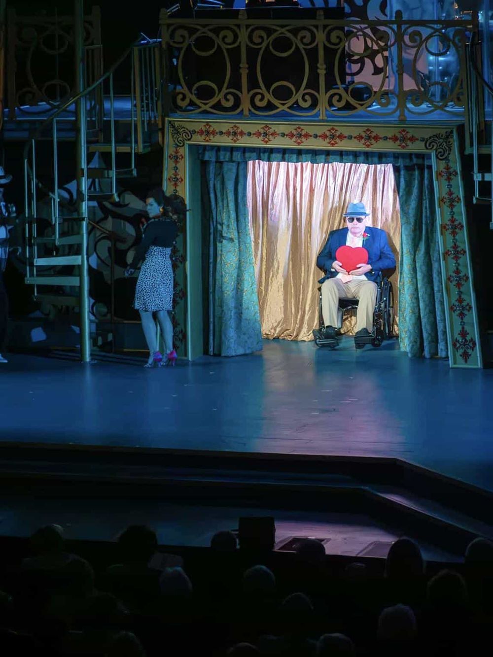 Colorful theatrical scene with a man in wheelchair holding a heart-shaped prop on stage.