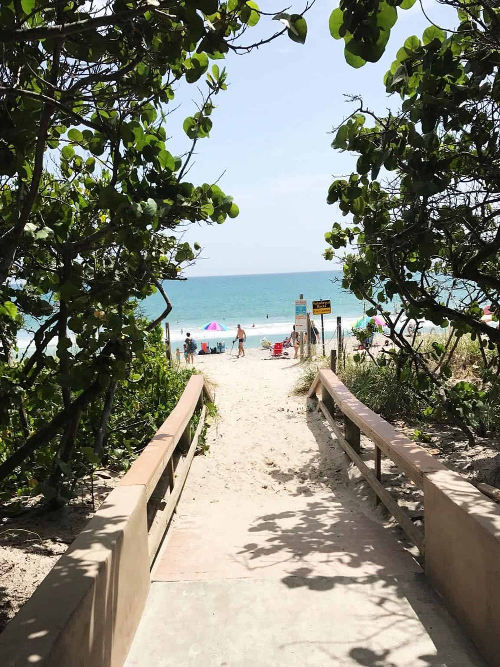 Beach access pathway leading to vibrant sunlit shoreline with umbrellas and people enjoying the ocean view.