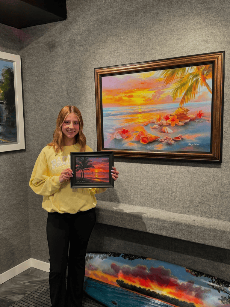 Vibrant coastal sunset painting displayed in an art gallery with a woman holding a framed sunset photo.