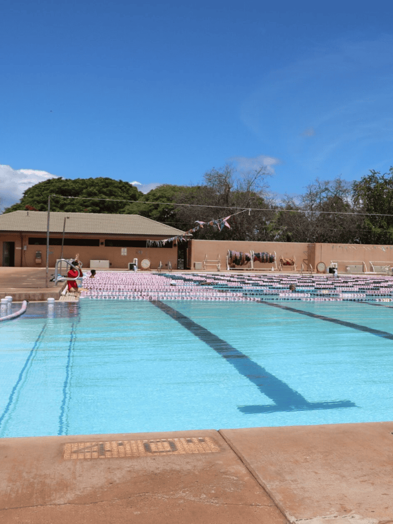Outdoor public swimming pool with lanes, poolside lounge chairs, and diving board under clear blue sky.
