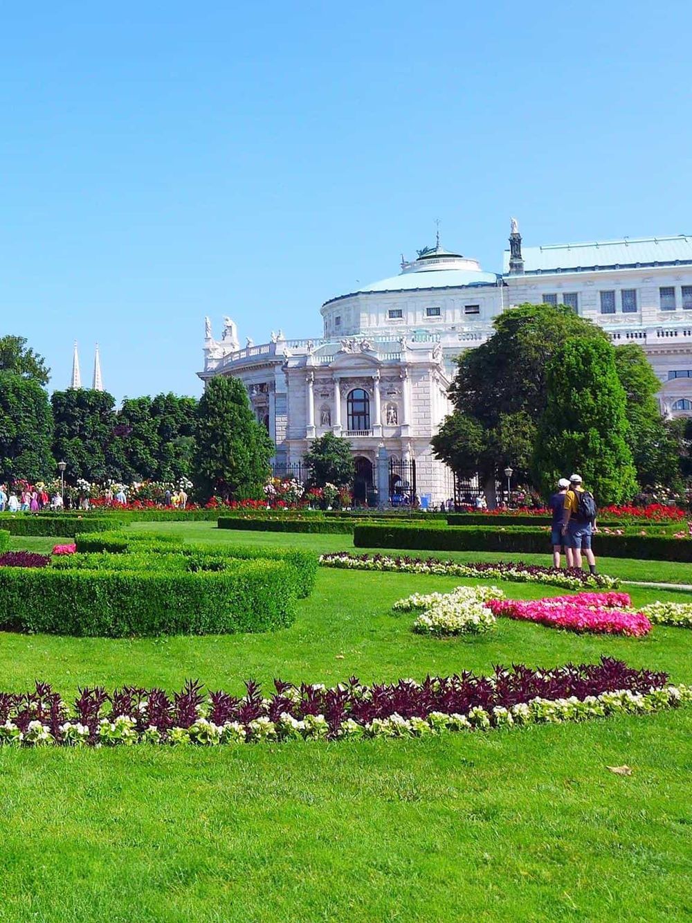 Colorful flower garden and historic white building with domed roof, featuring lush greenery and visitors in Vienna.