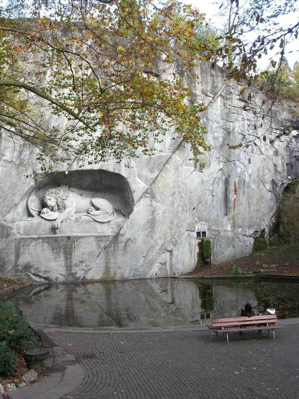 Lion carving in rock alcove at Quest for Directions, scenic outdoor location, nature-oriented park, historical monument, outdoor sightseeing, reflecting pond, autumn foliage.