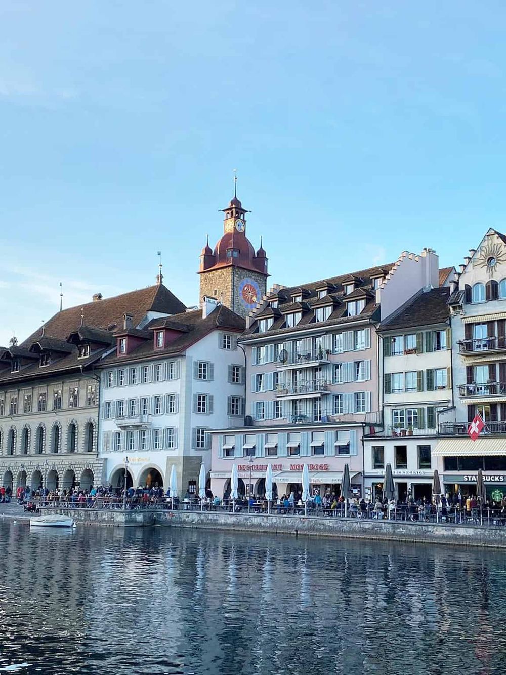 Colorful lakeside buildings in Switzerland with historic clock tower and outdoor cafes, picturesque mountain town scenery.
