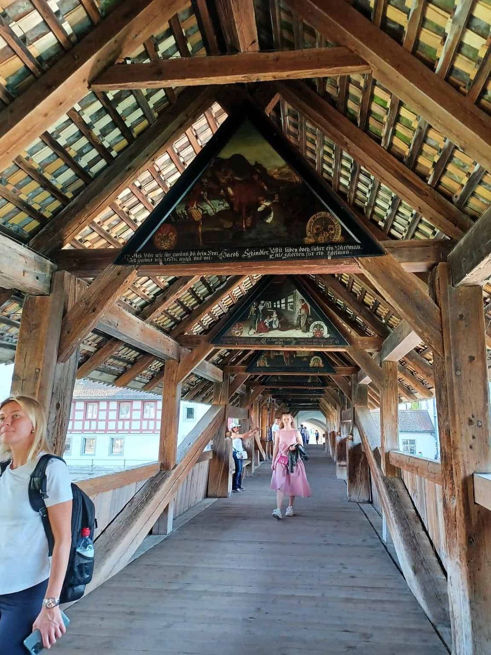 Historic covered wooden bridge with paintings, medieval architecture, and visitors exploring.