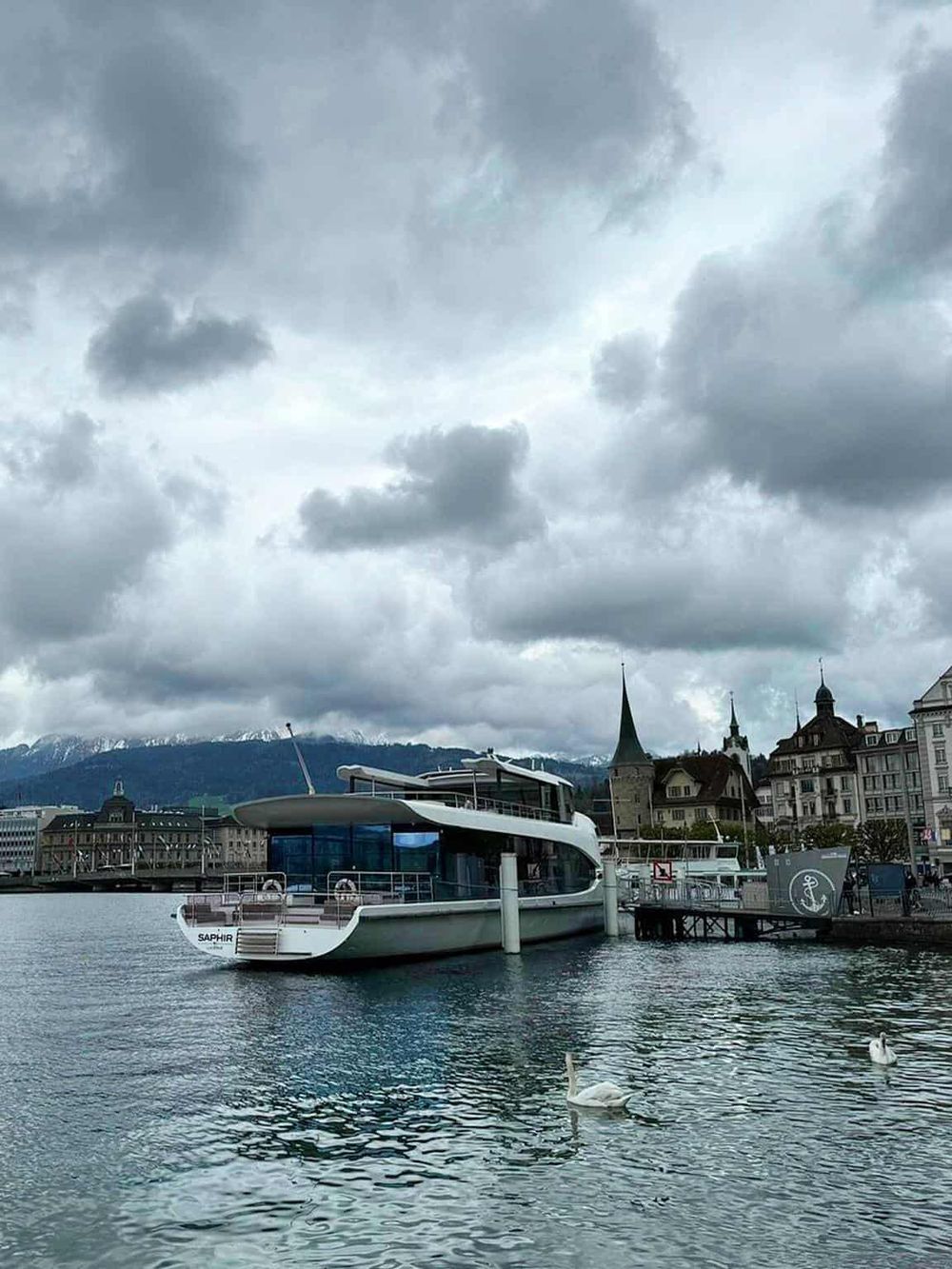 Modern luxury yacht docked at a scenic harbor with historic buildings and snow-capped mountains in the background.