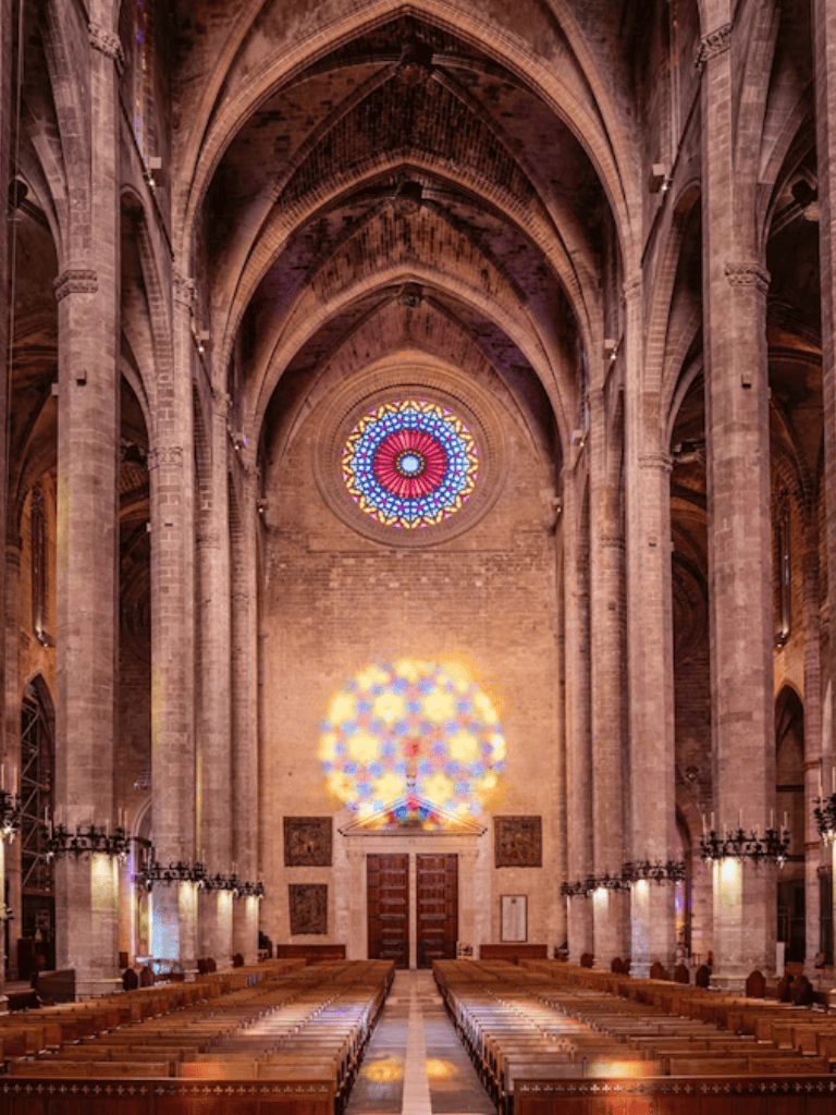 Ornate church interior with stained glass windows, soaring arches, and patterned light reflections, showcasing Gothic architecture.
