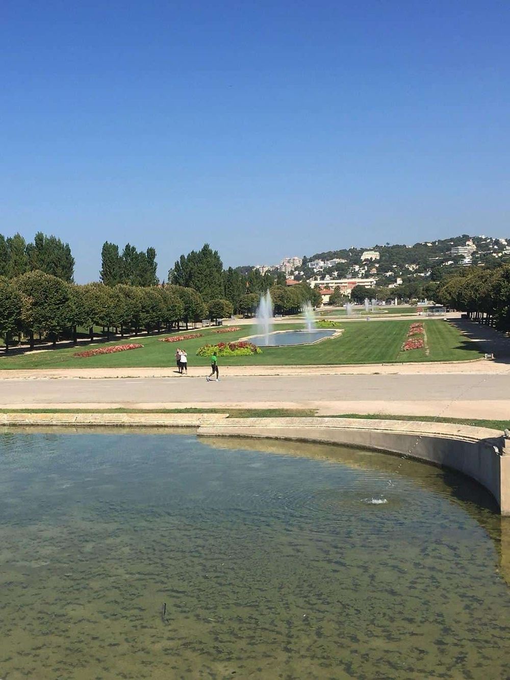 Green park with fountains and walking paths under clear blue sky, ideal for relaxation and outdoor activities.