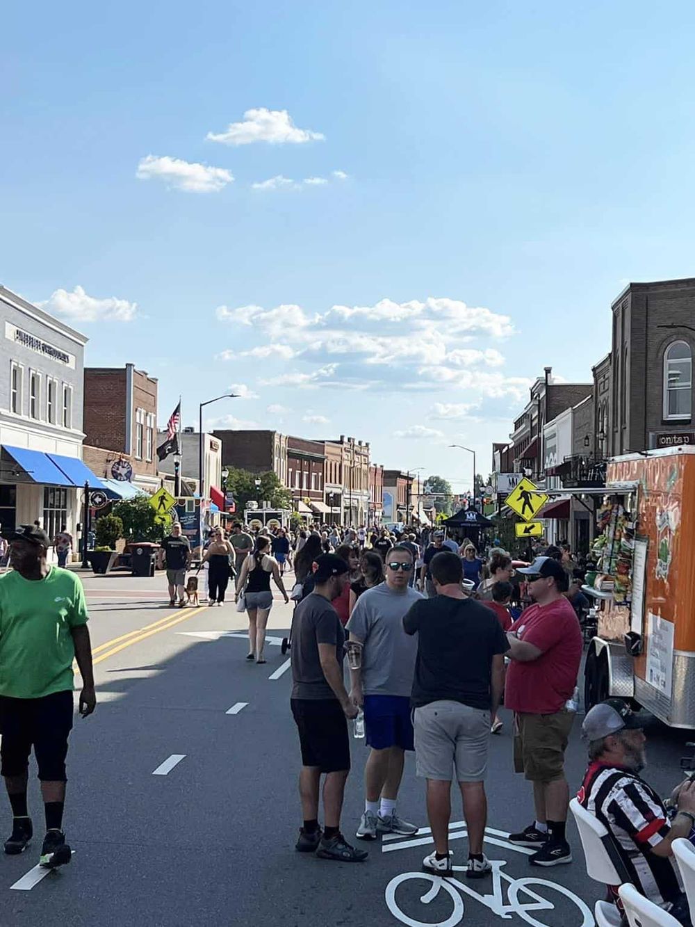 Vibrant downtown street scene with bustling crowd, shops, and food trucks on a sunny day.