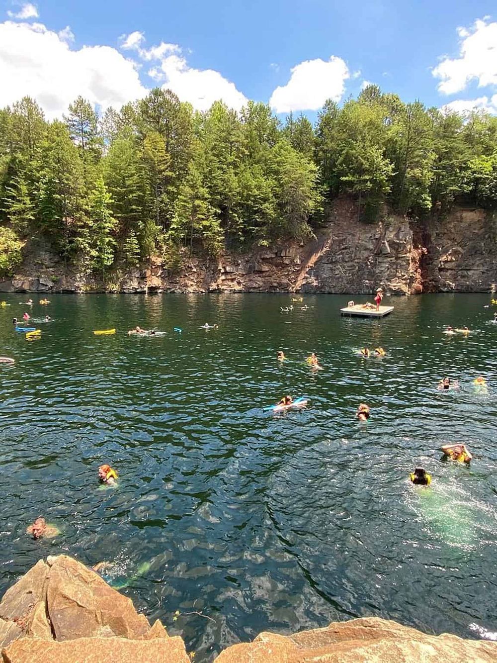 Swimmers enjoying a scenic outdoor lake with clear water surrounded by lush green trees and rocky cliffs.