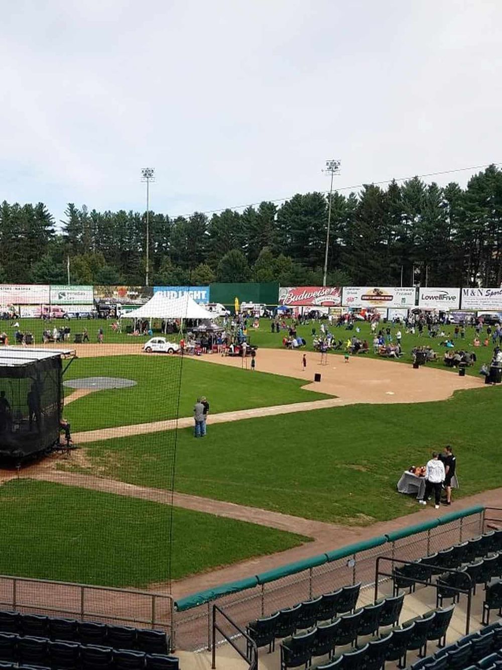 Open-air baseball stadium with spectators, tents, and advertising banners for local brands.