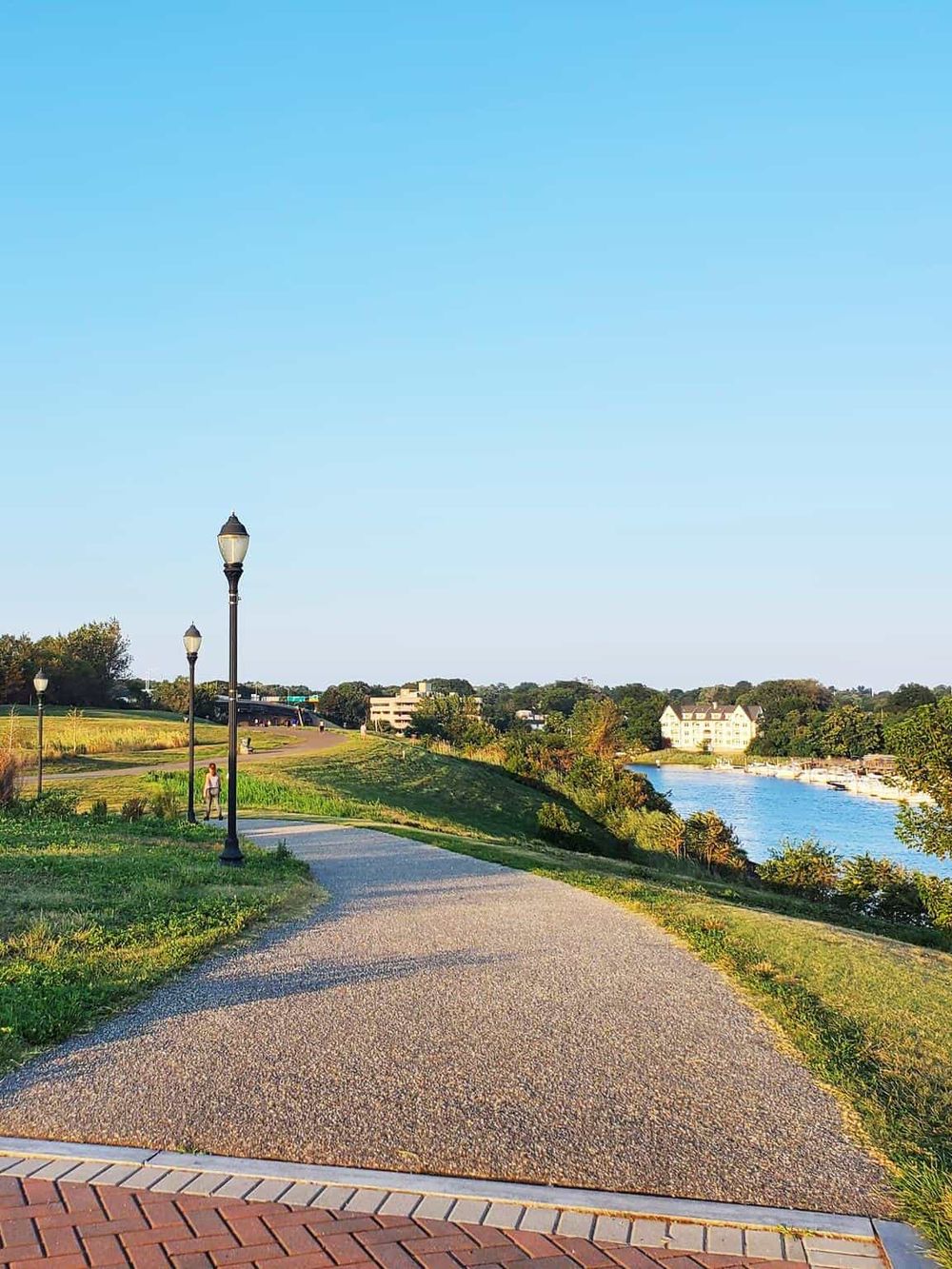 Scenic riverside park with walking path, lampposts, and lush greenery under a bright blue sky.
