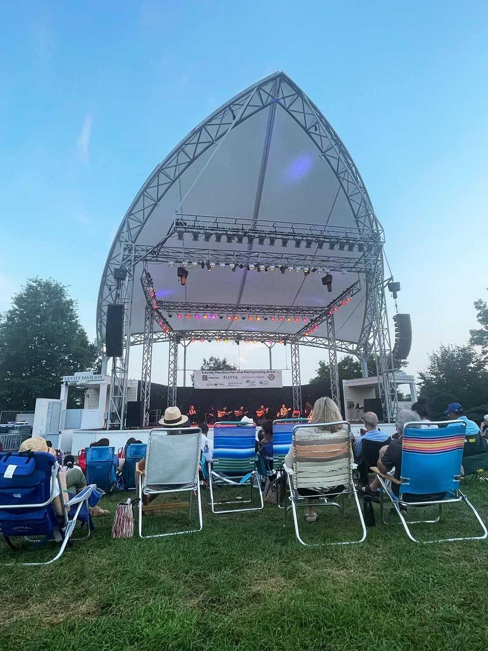 Open-air stage at outdoor music festival with audience, blue sky, and green trees in background.