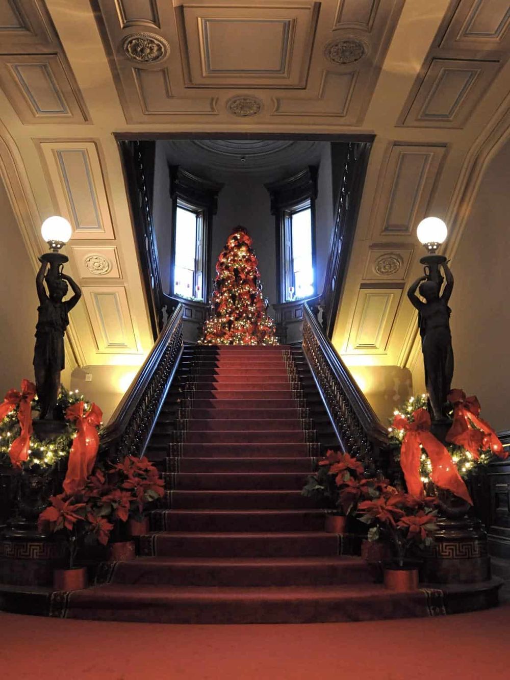 Intricate staircase decorated for Christmas with poinsettias and lights, leading to a grand tree flanked by statues.