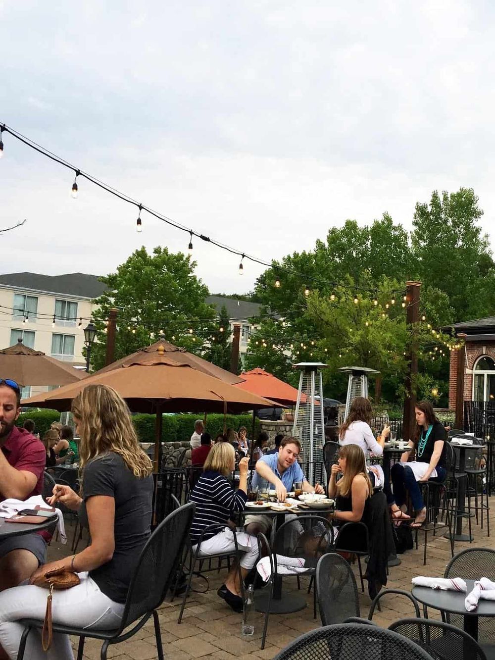Outdoor dining at a restaurant with string lights, patio umbrellas, and lush greenery.