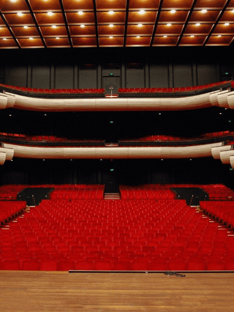Bright red theater seats in a large auditorium with tiered balconies and a wooden stage.
