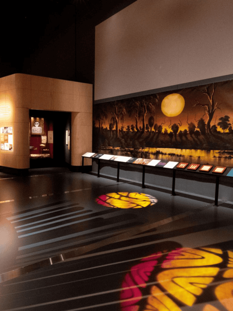 Vivid museum exhibit with butterfly shadow projections on the floor and large artwork of a sunset landscape.
