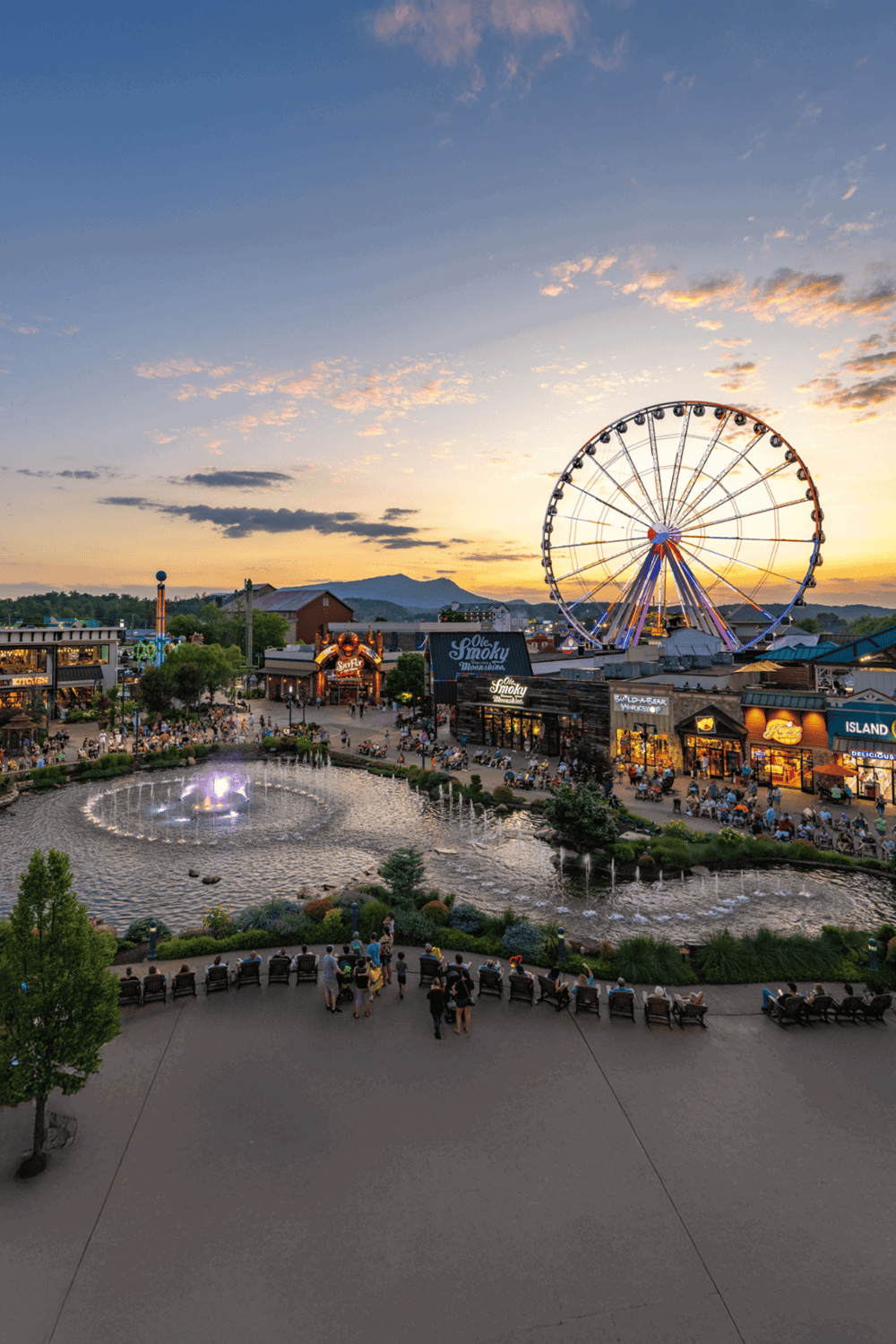 Amusement park at sunset with Ferris wheel, fountains, and visitors enjoying evening fun.