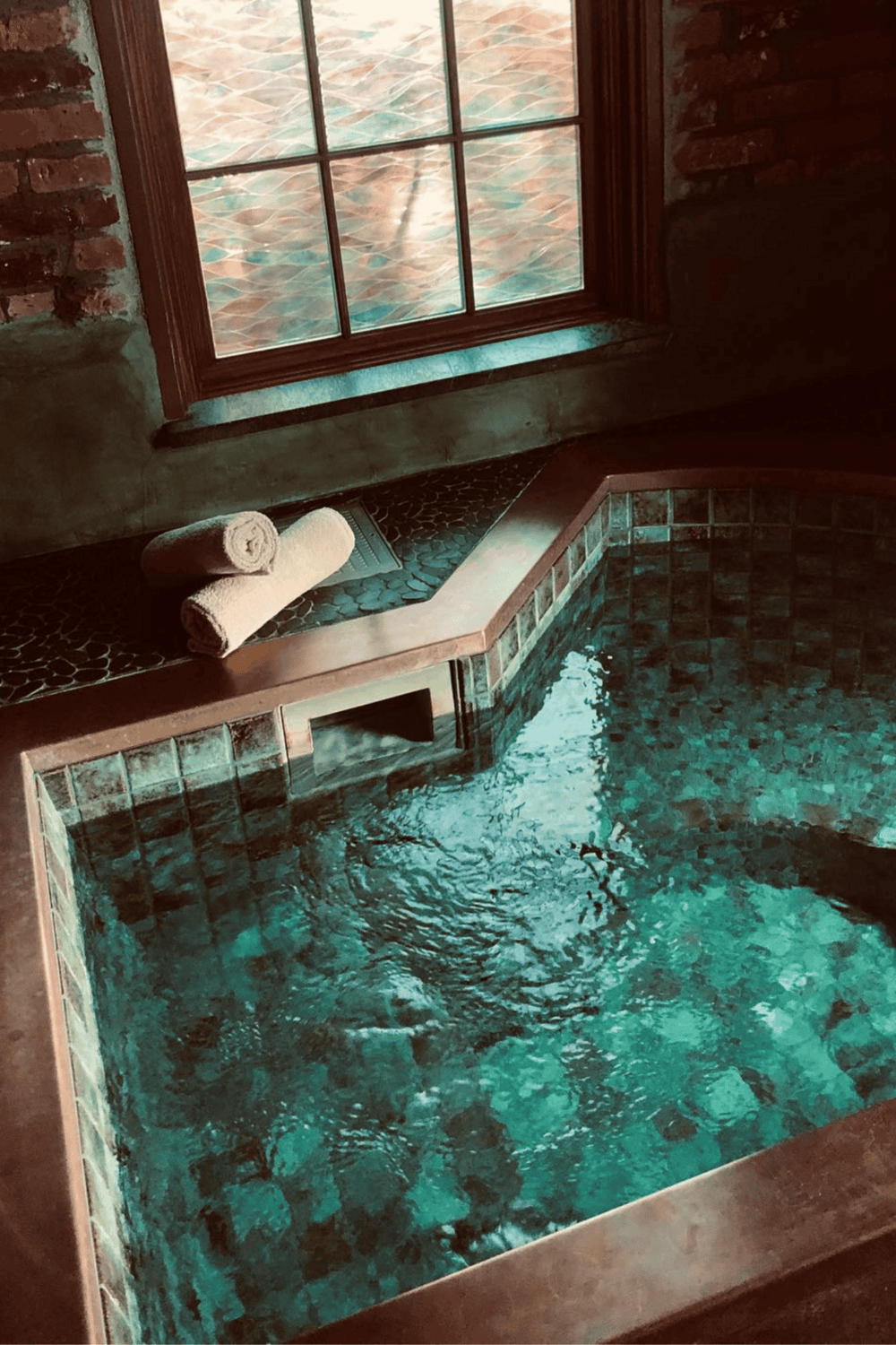 Soaking tub with brick accents and rolled towels near a window, creating a serene relaxation space.