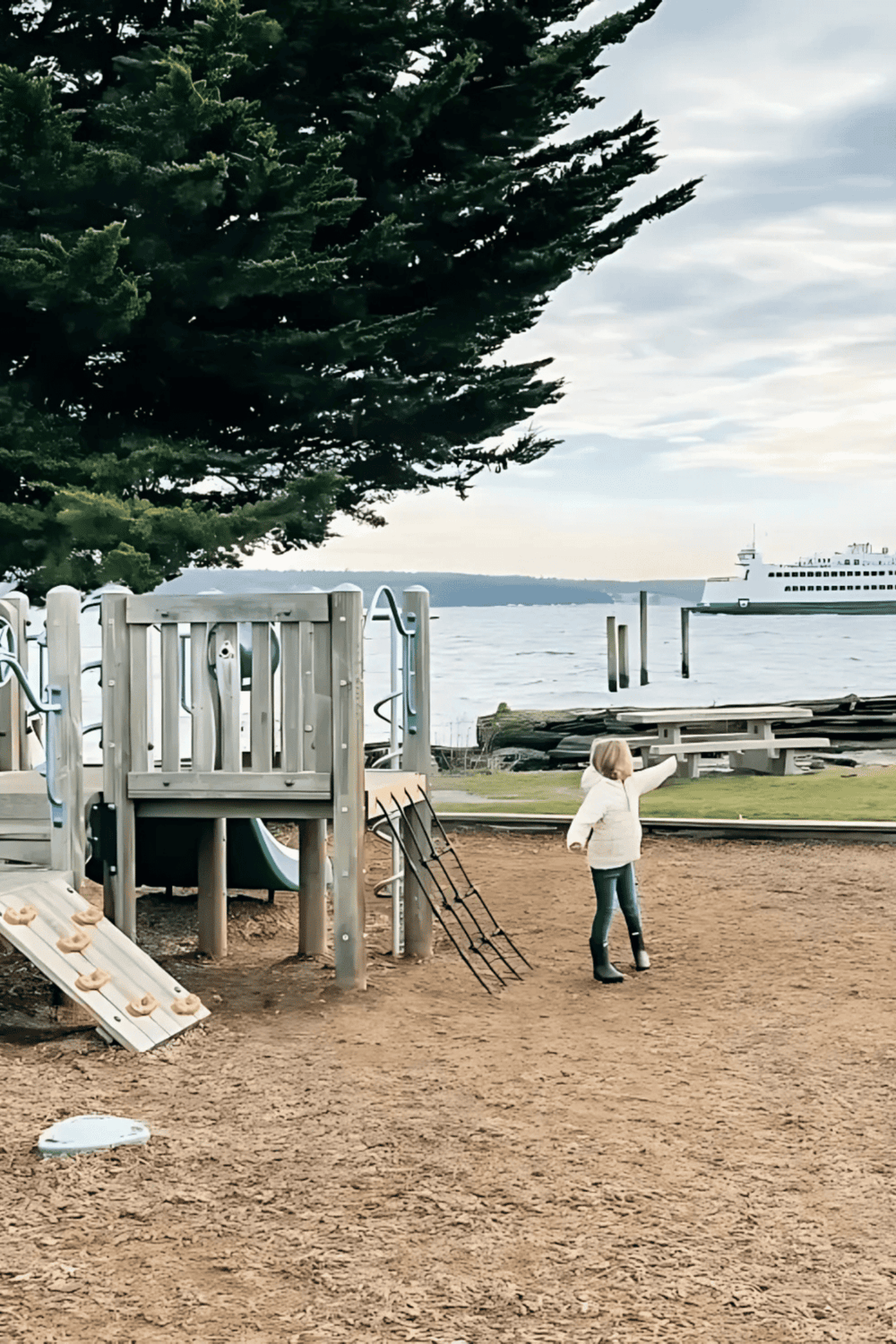 Child playing at a waterfront park with playground equipment and a ferry in the background.