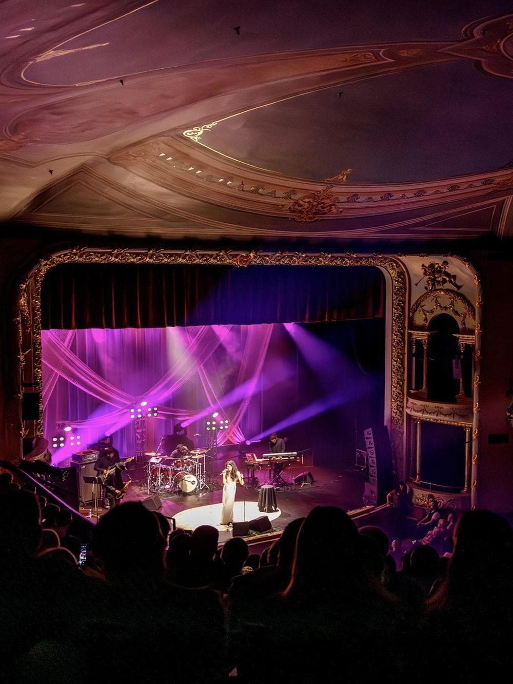 Performing band live music concert in a historic theater with ornate ceiling and elegant gold detailing.