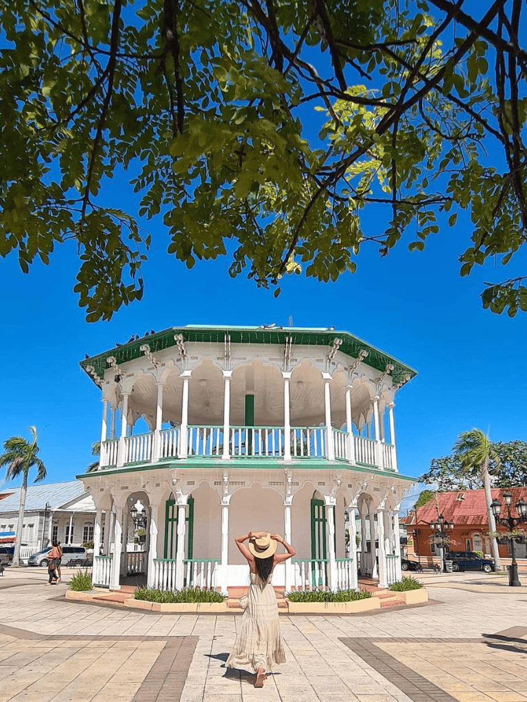Historic gazebo in a vibrant park with clear blue sky, palm trees, and a woman enjoying the sunny day.
