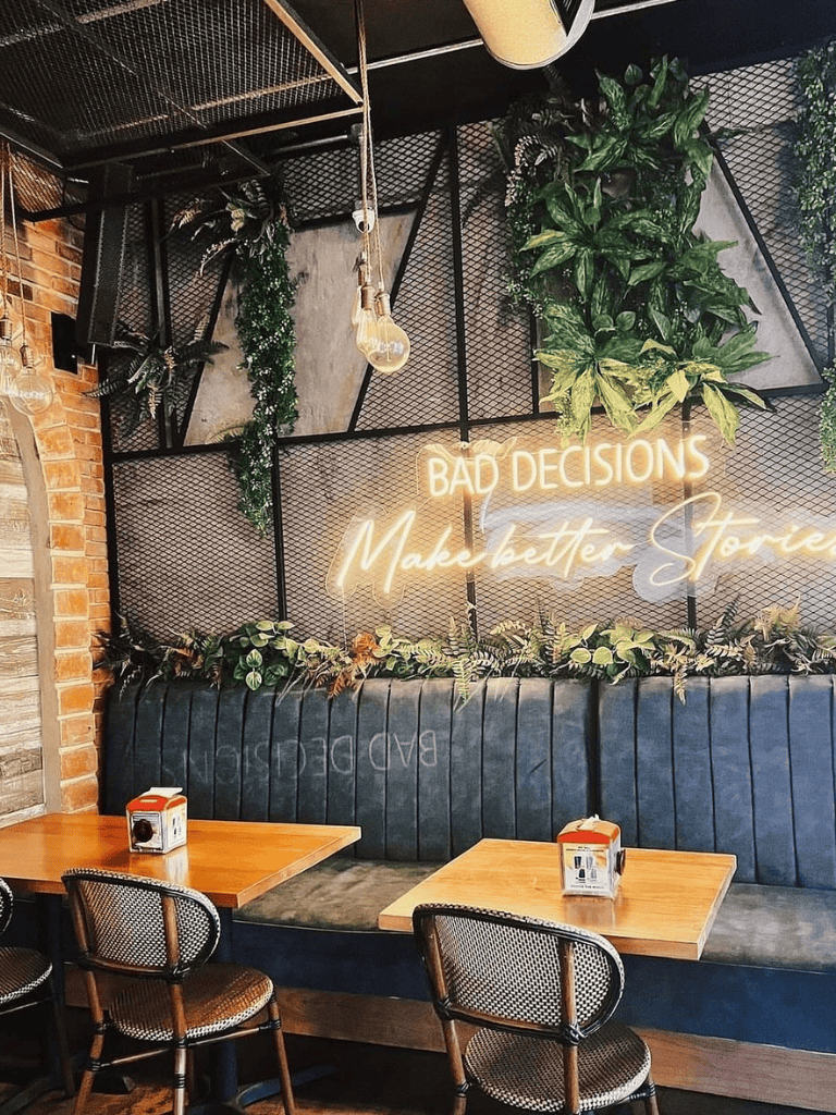 Modern urban café interior with neon "Bad Decisions Make Better Stories" sign and lush greenery.