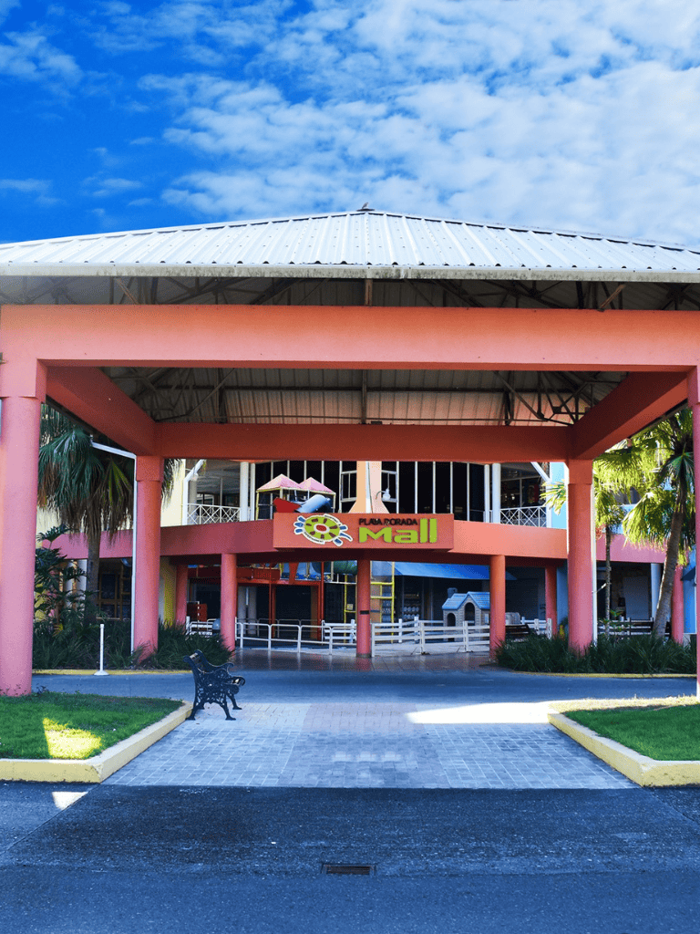 Bright pink mall entrance with a curved roof at Plaza Forada Mall, Florida, under blue skies, with green landscaping.