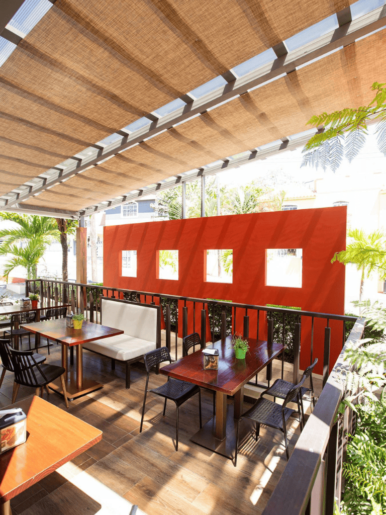 Bright outdoor restaurant terrace with wooden tables, black chairs, tropical plants, and shaded sunroof.