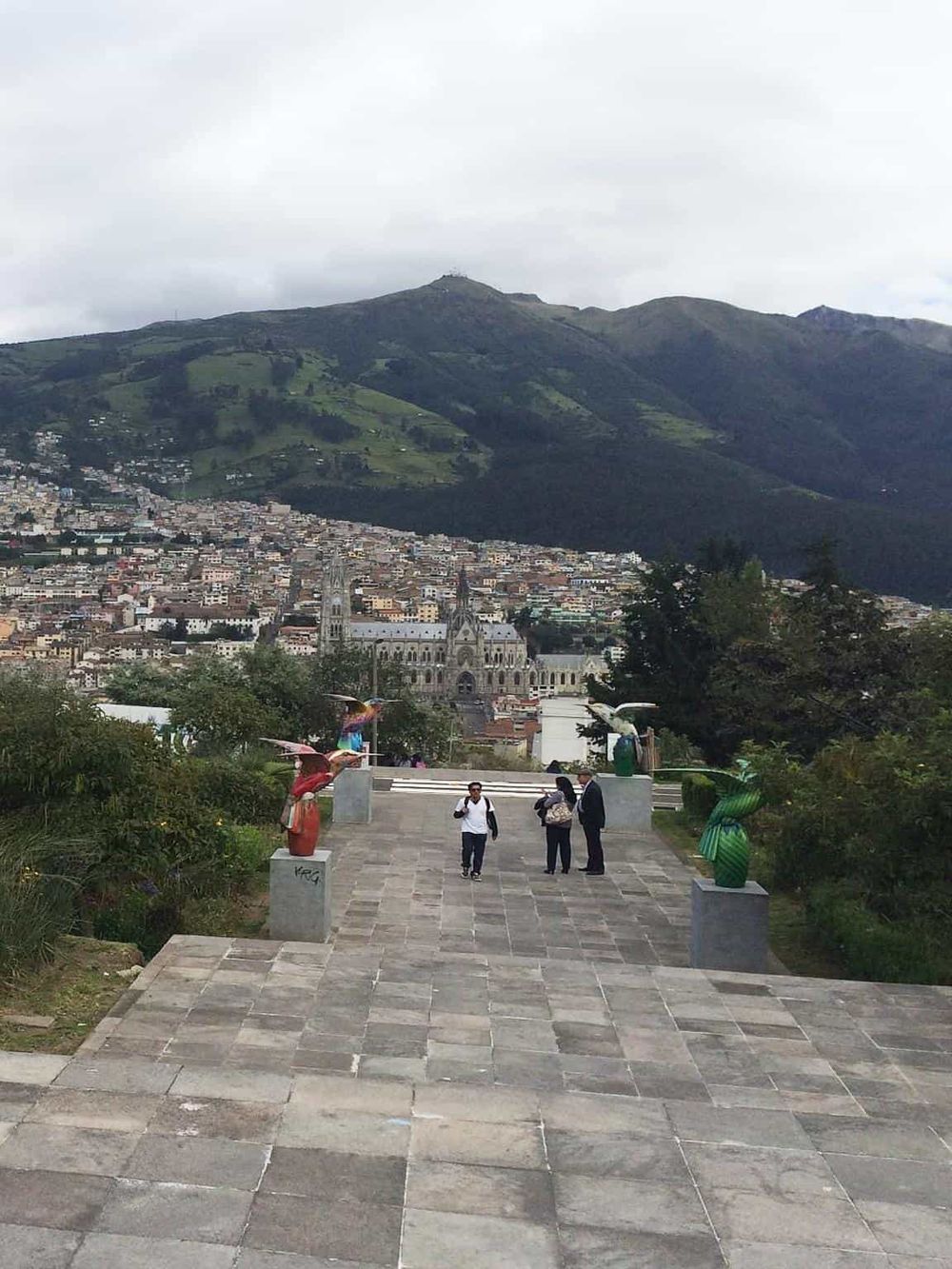 Colorful bird sculptures overlooking a city and lush mountain in Ecuador, popular tourist attraction.