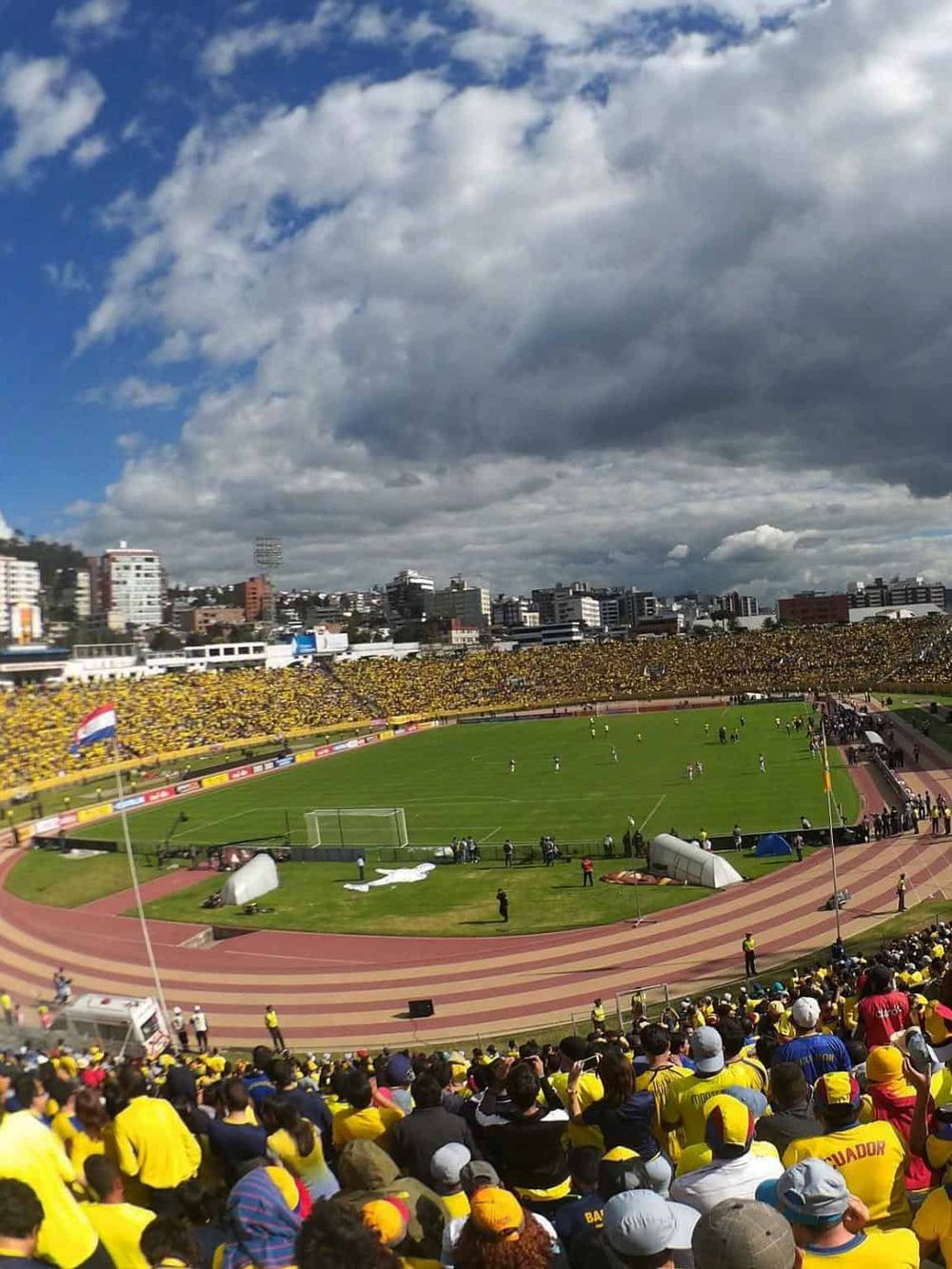 Vibrant soccer stadium filled with enthusiastic fans wearing yellow, overlooking a lively match on a cloudy day.