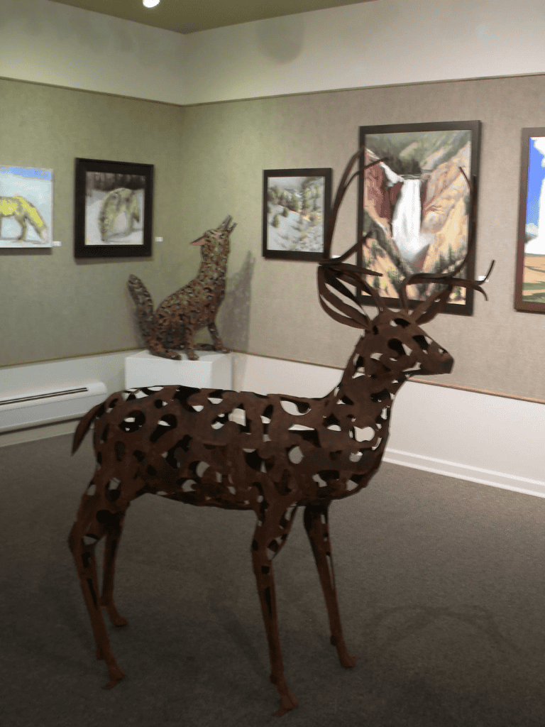 Intricate metal deer sculpture displayed in an art gallery with paintings backdrop.