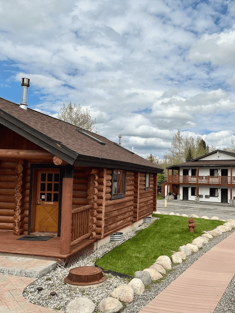 Charming log cabin exterior with landscaped yard and modern building in the background - ideal for travel planning.