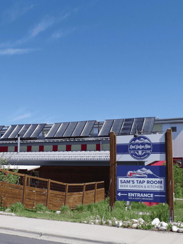 Blue sky with solar panel-covered building, rustic wooden fence, and QuestForDirections sign for Sam's Tap Room and beer garden.
