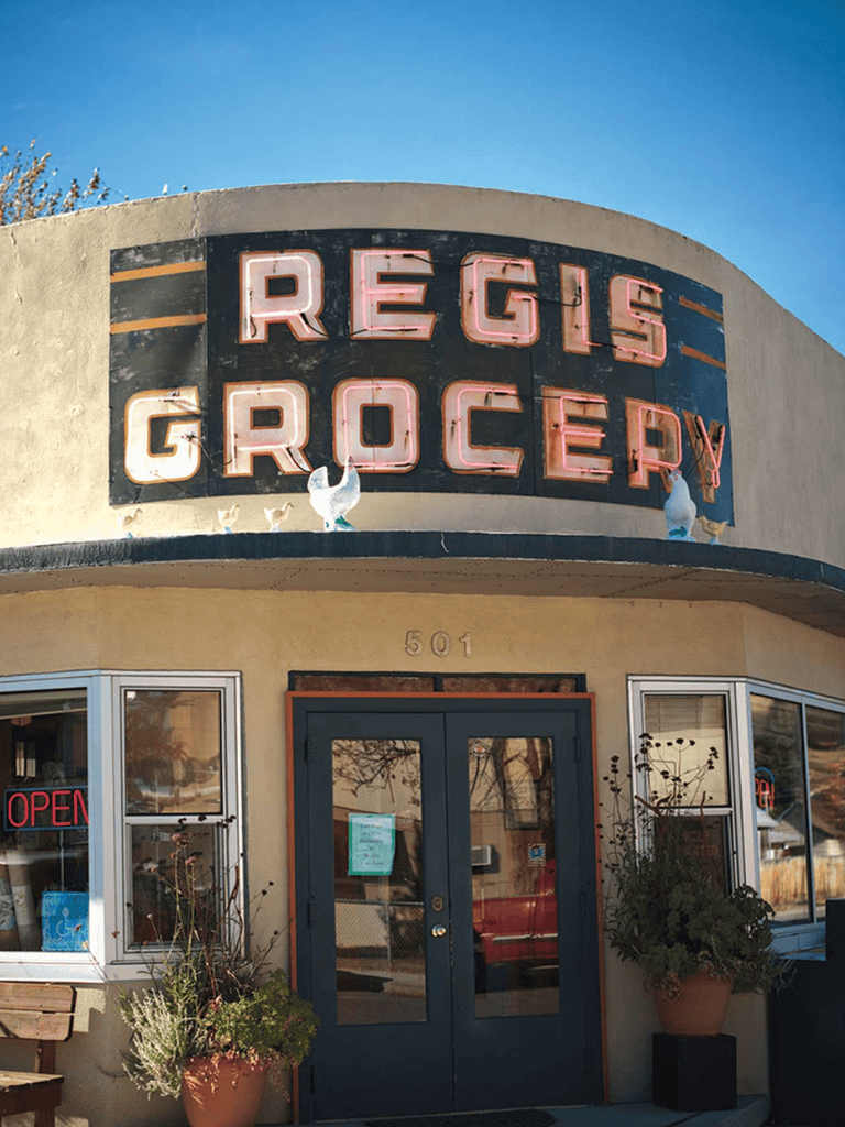 Neighborhood grocery store with vintage neon sign, inviting storefront with potted plants, perfect for local shopping and community access.