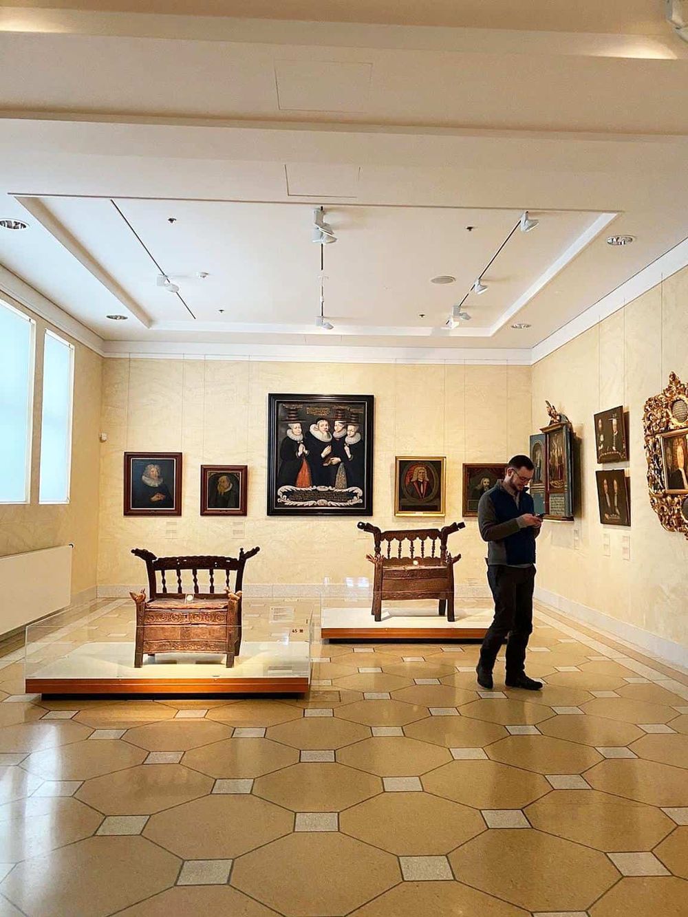 Colorful art gallery with framed paintings, wooden artifacts, and a visitor using a smartphone in the museum.