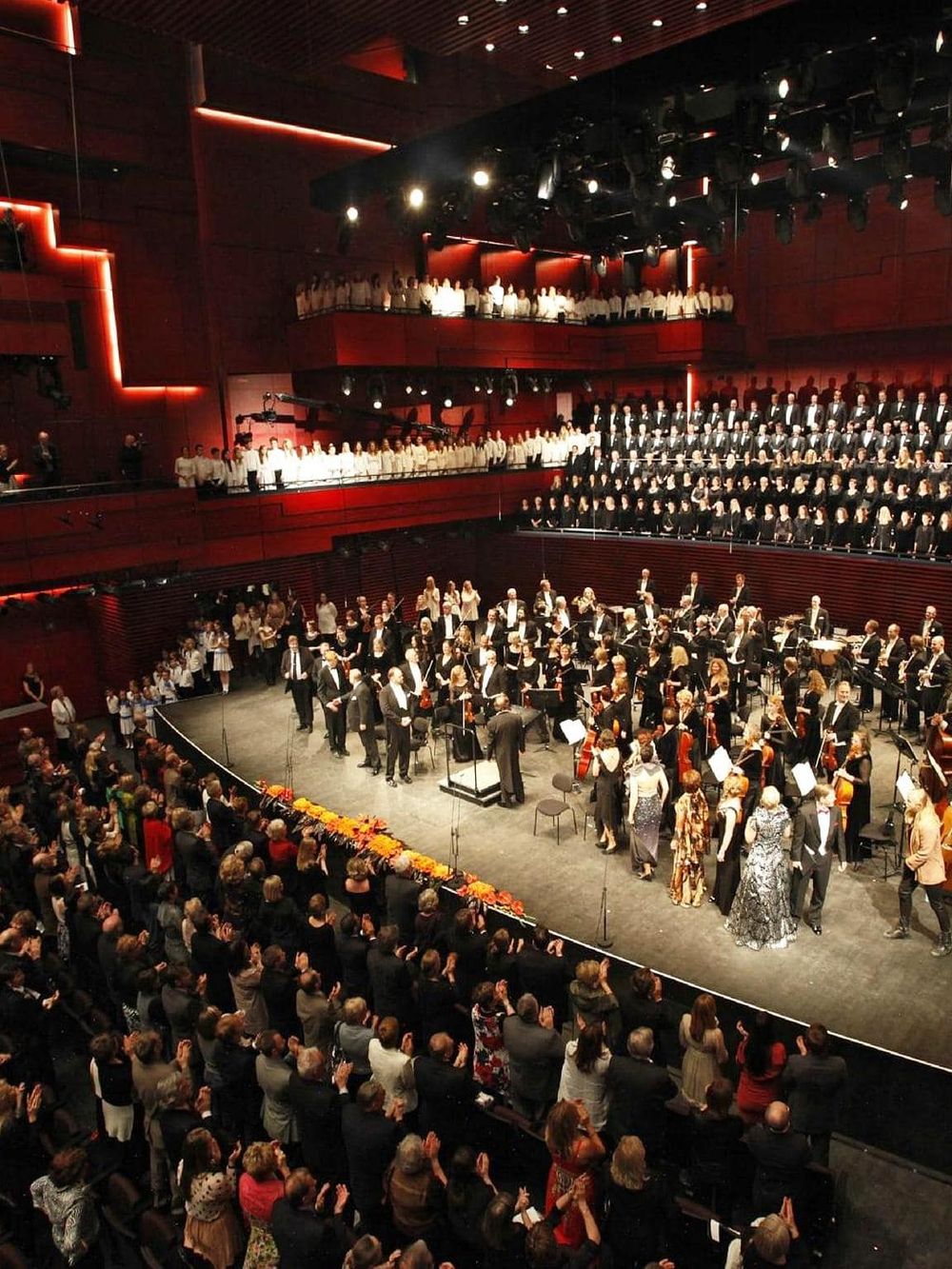 A large orchestral performance in a modern concert hall with an engaged audience and choir on upper levels.