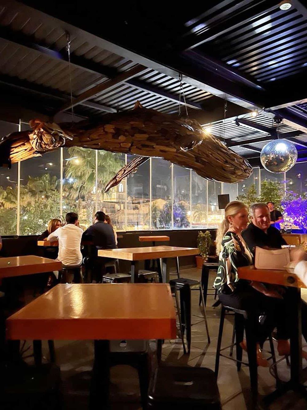 Whale art installation hanging from ceiling at Quest For Directions restaurant, modern ambiance, people dining, night scene.