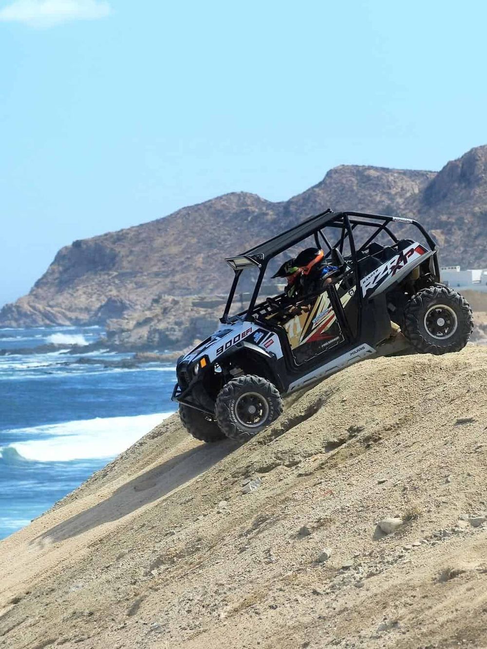 Off-road utility terrain vehicle (UTV) driving on sandy beach with rugged cliffs and ocean in background.